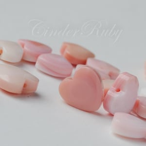 May include: A collection of pink heart-shaped shell beads with a hole for stringing. The beads are smooth and have a natural, iridescent sheen.