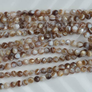 May include: A collection of round, polished beads arranged in multiple horizontal strands. Each bead features a swirling pattern of brown, tan, and cream colors with a subtle iridescent sheen, creating a natural, banded appearance across the surface.