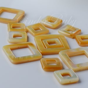 May include: A collection of 12 yellow and white shell square-shaped beads with a hole in the center. The beads are arranged in a random pattern on a white surface.