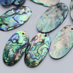 May include: A collection of oval-shaped abalone shell pendants featuring iridescent surfaces with swirling patterns of blue, green, and purple. Each flat, polished shell piece includes a small pre-drilled hole at the top for hanging or attachment.