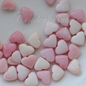 May include: A collection of pink and white heart-shaped beads made of mother of pearl. The beads are arranged on a white surface.