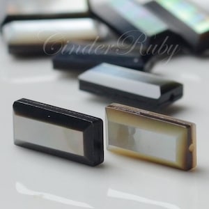 以下が含まれることがあります： 黒い縁取りのある長方形の虹色の真珠貝のカボションが2つ。カボションの長さは約2.5cm、幅は約1.25cmです。