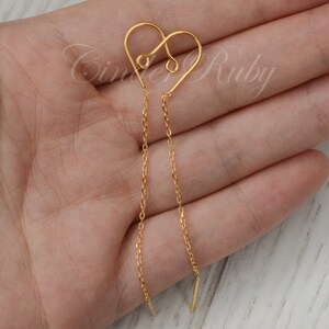 May include: Gold heart shaped dangle earrings with a delicate chain. The earrings are shown on a hand.