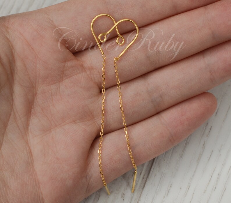 May include: Gold heart shaped dangle earrings with a delicate chain. The earrings are shown on a hand.