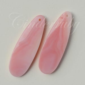 May include: Two elongated, teardrop-shaped pendants in a soft, pale pink hue with a smooth, marbled texture. Each piece features a small, circular hole at the top for suspension, showcasing a consistent, polished finish throughout the surface.