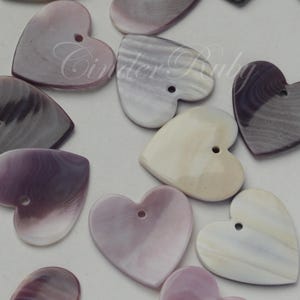 May include: A collection of heart-shaped buttons in shades of purple, white, and gray. Each button has a hole for sewing and a glossy finish. The buttons vary in color and pattern, creating a unique assortment for crafts and sewing projects.