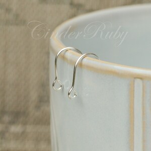 May include: Two silver earring hooks, also known as ear wires, are displayed on a white ceramic surface. The hooks are made of a thin, smooth metal and have a simple, elegant design.