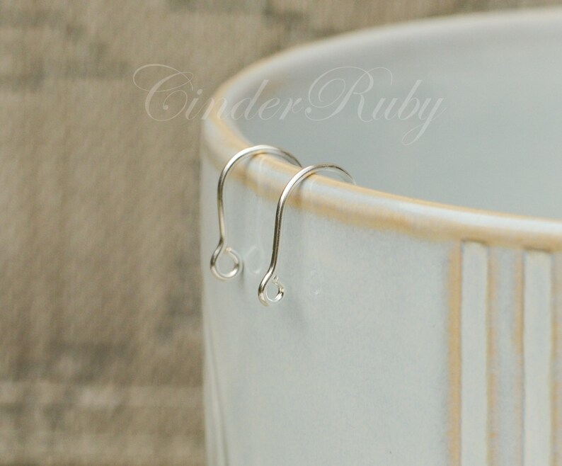May include: Two silver earring hooks, also known as ear wires, are displayed on a white ceramic surface. The hooks are made of a thin, smooth metal and have a simple, elegant design.