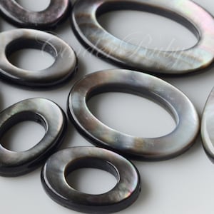 May include: A collection of oval-shaped, black mother-of-pearl shell beads with a hole in the center. The beads have a smooth, polished surface and a subtle iridescent sheen.