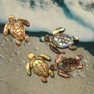 May include: Four decorative turtle figurines with iridescent shells and gold-toned accents are arranged on a sandy surface near the ocean. The turtles have detailed shell patterns and flipper-like appendages, creating a coastal theme.