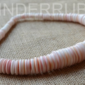 May include: A necklace made of round, flat, pink and white shell beads. The beads are strung together to form a circular shape. The necklace is displayed on a tan surface.