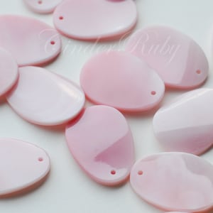 May include: A collection of pink oval-shaped shell beads with a hole drilled through the top. The beads are smooth and have a pearlescent finish.