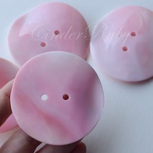 May include: Close-up of several large, round, pink buttons with two holes each. The buttons have a swirled, marbled effect with lighter pink and white tones. The buttons are on a white background.
