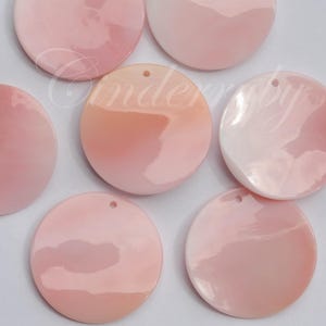 May include: A collection of round, pink shell pendants with a glossy finish. Each pendant has a small hole at the top for stringing. The pendants vary in shades of pink, from light to a deeper rose color.