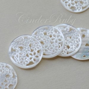 May include: Five white, round, decorative buttons with a floral design. The buttons are made of a pearlescent material and have a delicate, intricate pattern.