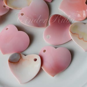 May include: A collection of pink and white heart-shaped shell charms with a hole at the top for hanging. The shells have a natural, iridescent finish.