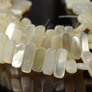 May include: Close-up of a strand of rectangular, off-white mother-of-pearl beads. The beads have a lustrous, iridescent sheen, reflecting light with hints of pink, green, and yellow. The beads are strung together, creating a textured, organic pattern.