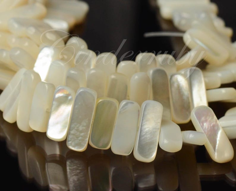 May include: Close-up of a strand of rectangular, off-white mother-of-pearl beads. The beads have a lustrous, iridescent sheen, reflecting light with hints of pink, green, and yellow. The beads are strung together, creating a textured, organic pattern.