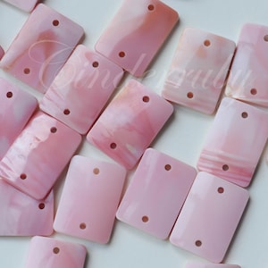 Könnte beinhalten: Eine Sammlung rechteckiger, rosa Perlen mit jeweils zwei Löchern. Die Perlen haben eine glänzende Oberfläche und weisen Muster aus helleren rosa und weißen Marmorierungen auf. Geeignet für die Schmuckherstellung.