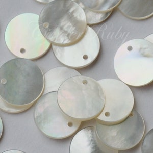 May include: A collection of white, round, iridescent mother of pearl shell discs with a hole in the center. The discs are scattered on a white surface.