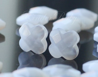 May include: Close-up of several white, four-petal flower-shaped beads. The beads have a faceted surface, reflecting light. The background is blurred, with other similar beads visible. These beads are suitable for jewelry making.