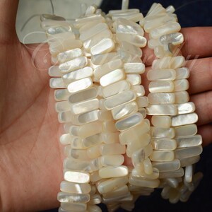 May include: Close-up of several strands of rectangular, off-white mother-of-pearl beads. The beads are strung together, creating a textured, organic look. The beads have a slight iridescent sheen, reflecting light. The overall theme is jewelry.