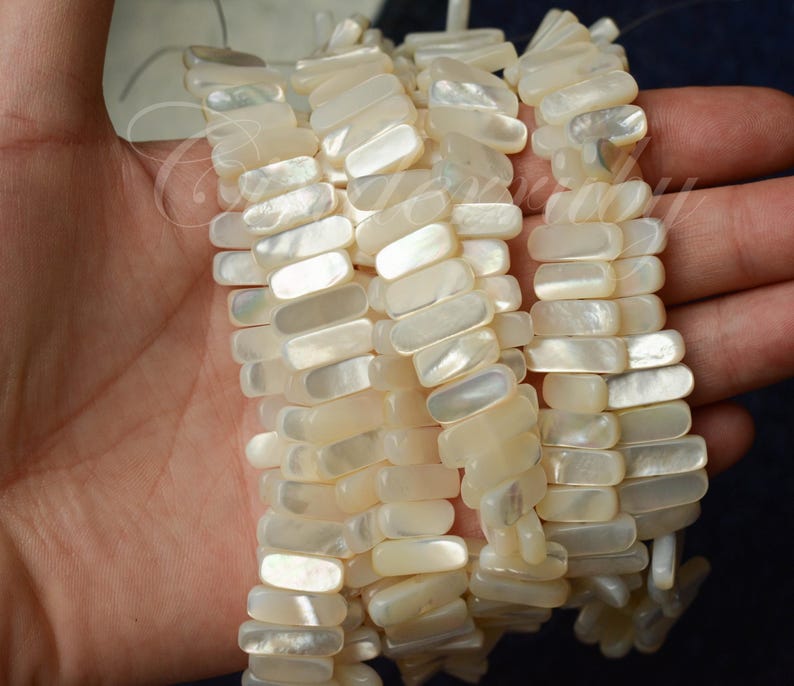 May include: Close-up of several strands of rectangular, off-white mother-of-pearl beads. The beads are strung together, creating a textured, organic look. The beads have a slight iridescent sheen, reflecting light. The overall theme is jewelry.
