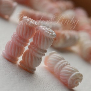 May include: Close-up of pink and white shell beads with a twisted design. The beads are arranged in a pile on a white surface.