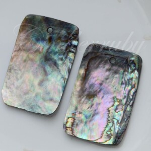 May include: Two rectangular abalone shell pendants featuring an iridescent surface with swirling hues of blue, green, purple, and pink. Each piece has a single circular hole drilled near the top edge for suspension, showcasing a natural finish.