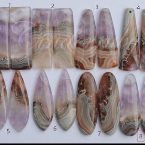 May include: A collection of sixteen polished gemstone pendants arranged in eight numbered pairs. The stones feature banded patterns in shades of purple, white, brown, and gray, with various shapes including rectangles, teardrops, and ovals.
