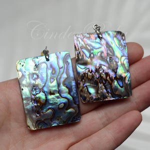 May include: A pair of iridescent abalone shell earrings with silver hooks. The earrings are rectangular in shape and have a natural, organic pattern.