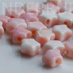 May include: A collection of star-shaped beads in shades of pink and white. Each bead has a hole for stringing. The beads have a glossy finish and are scattered on a white surface.