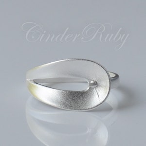 May include: A silver ring with a unique, open design. The ring features a smooth, oval-shaped top with a small, round accent. The ring is made of a shiny, metallic material.