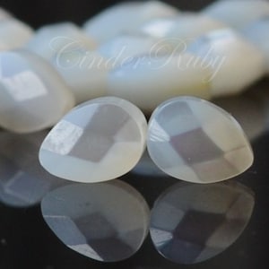 May include: Two white iridescent gemstone cabochons with a geometric pattern in shades of gray and purple. The cabochons are shaped like teardrops and have a smooth, polished surface.
