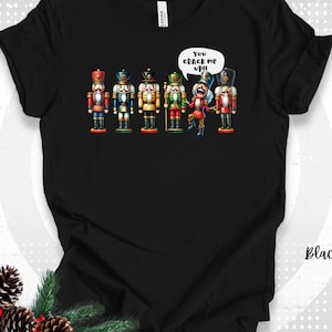 May include: Black t-shirt featuring a colorful illustration of six nutcracker soldiers. One nutcracker has a speech bubble that says "You crack me up!!" The word "Black" is printed on the shirt.