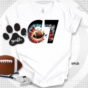 Puede incluir: Camiseta blanca con un gráfico temático de fútbol. El diseño presenta el número "67" con un balón de fútbol en el centro, rodeado de un efecto de explosión. También hay una huella de pata negra con la palabra "youth". Un balón de fútbol y un silbato también son visibles.