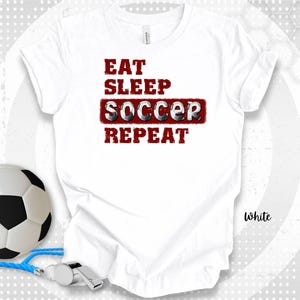 以下が含まれることがあります： 赤と白で「EAT SLEEP SOCCER REPEAT」の文字が入った白いTシャツ。サッカーボール、ホイッスル、青いランヤードが画像にあります。「White」の文字がシャツにプリントされています。