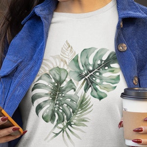 以下が含まれることがあります： 緑と茶色の熱帯の葉の水彩画のデザインが施された白いTシャツ