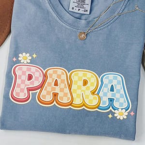 Może przedstawiać: Jasnoniebieska koszulka Comfort Colors z napisem "PARA" w stylu retro w szachownicę. Koszulka ma okrągły dekolt i kr&oacute;tkie rękawy. Złoty naszyjnik z wisiorkiem jest zarzucony na koszulkę. Koszulka jest złożona.