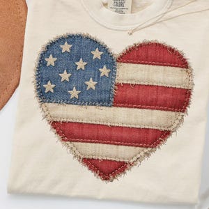 Może przedstawiać: Kremowy t-shirt z sercowatym wzorem przypominającym amerykańską flagę. Serce składa się z niebieskiego dżinsu z białymi gwiazdami oraz czerwono-białego paski. Koszulka ma okrągły dekolt i metkę z napisem "Comfort Colors".
