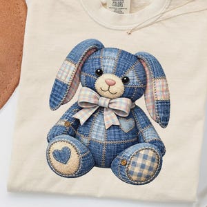 May include: Cream-coloured t-shirt featuring a denim patchwork bunny design with a pink bow and heart details. The bunny has long ears and button accents. The shirt has a Comfort Colors tag.