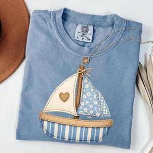 May include: A light blue Comfort Colors t-shirt with an appliqued sailboat design. The sailboat has a white sail with a brown heart, a blue and white floral sail, and a striped blue and white hull. A gold necklace is draped across the shirt.
