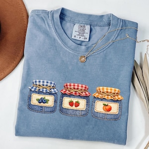 May include: A light blue Comfort Colors t-shirt featuring three embroidered jam jar designs. Each jar has a different fruit: blueberries, strawberries, and a peach. The jars have denim-like patches and are topped with gingham fabric lids. A gold necklace is draped across the shirt.