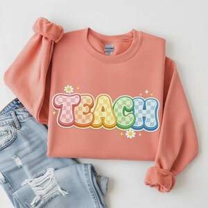 May include: A coral-colored sweatshirt with the word "TEACH" in a retro, colorful, checkered design. The letters are outlined in a bright pink, with a daisy and star accents. The sweatshirt is folded over a pair of blue jeans.