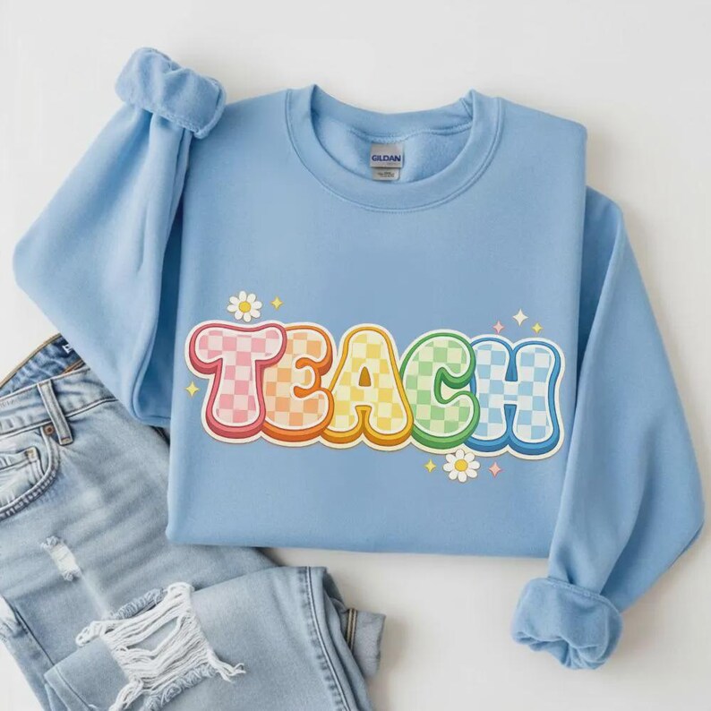 May include: A light blue sweatshirt with the word "TEACH" in a colorful, retro font. The letters are checkered in shades of pink, orange, yellow, green, and blue. Small white daisies and star accents surround the word. The sweatshirt is paired with blue jeans.