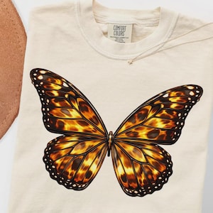 Op de afbeelding: Crèmekleurige Comfort Colors sweatshirt met een grote vlinderprint. De vlinder heeft gedetailleerde vleugels in bruin-, oranje- en zwarttinten. De sweatshirt heeft een ronde hals en een label met de tekst "Comfort Colors".