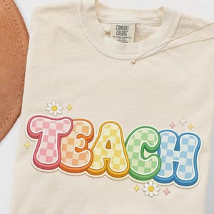 May include: Cream-colored t-shirt with the word "TEACH" in a colorful, retro font. The letters are outlined in white and filled with checkered patterns in various colors. Small daisy and star graphics are scattered around the word.