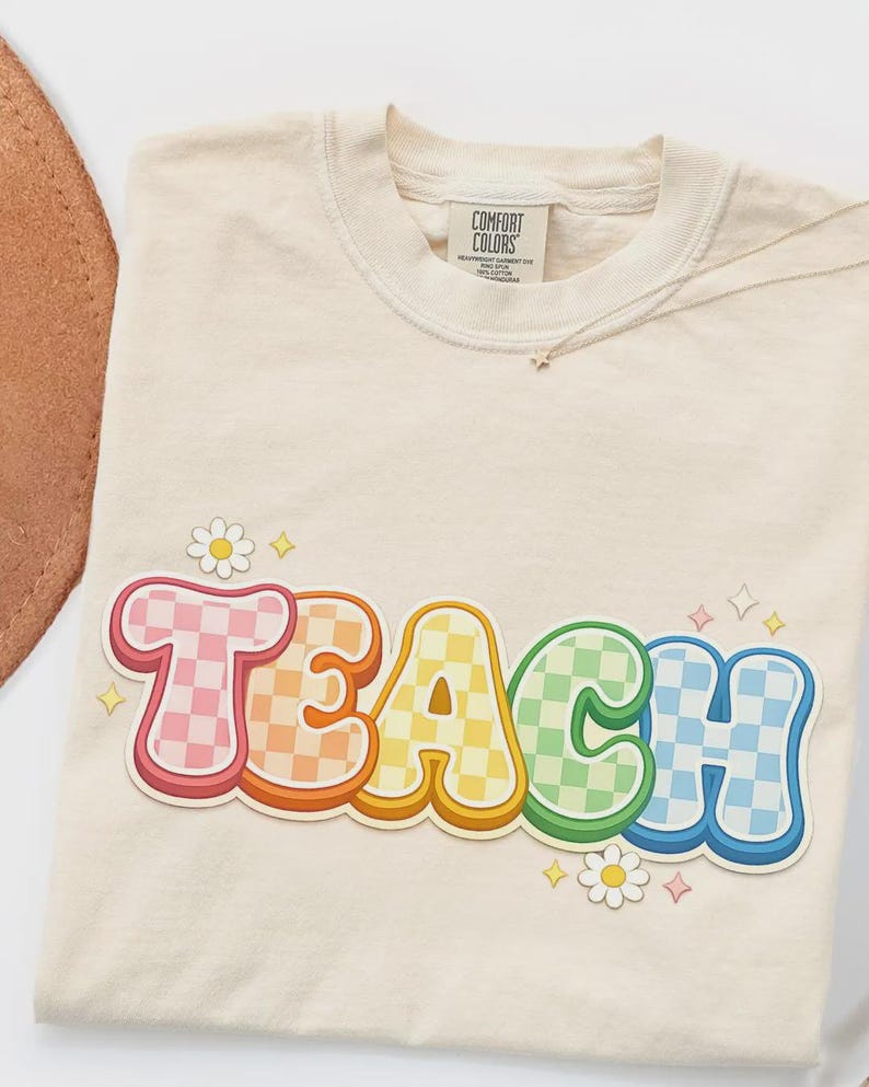 May include: Cream-colored t-shirt with the word "TEACH" in a colorful, retro font. The letters are outlined in white and filled with checkered patterns in various colors. Small daisy and star graphics are scattered around the word.