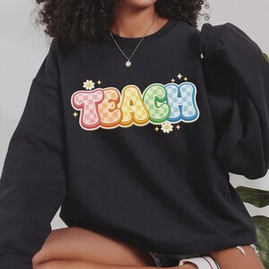 May include: Black sweatshirt with the word "TEACH" in a colorful, retro font. The letters are outlined in white and have a checkered pattern in shades of pink, orange, yellow, green, and blue. Small white flowers and stars accent the design.