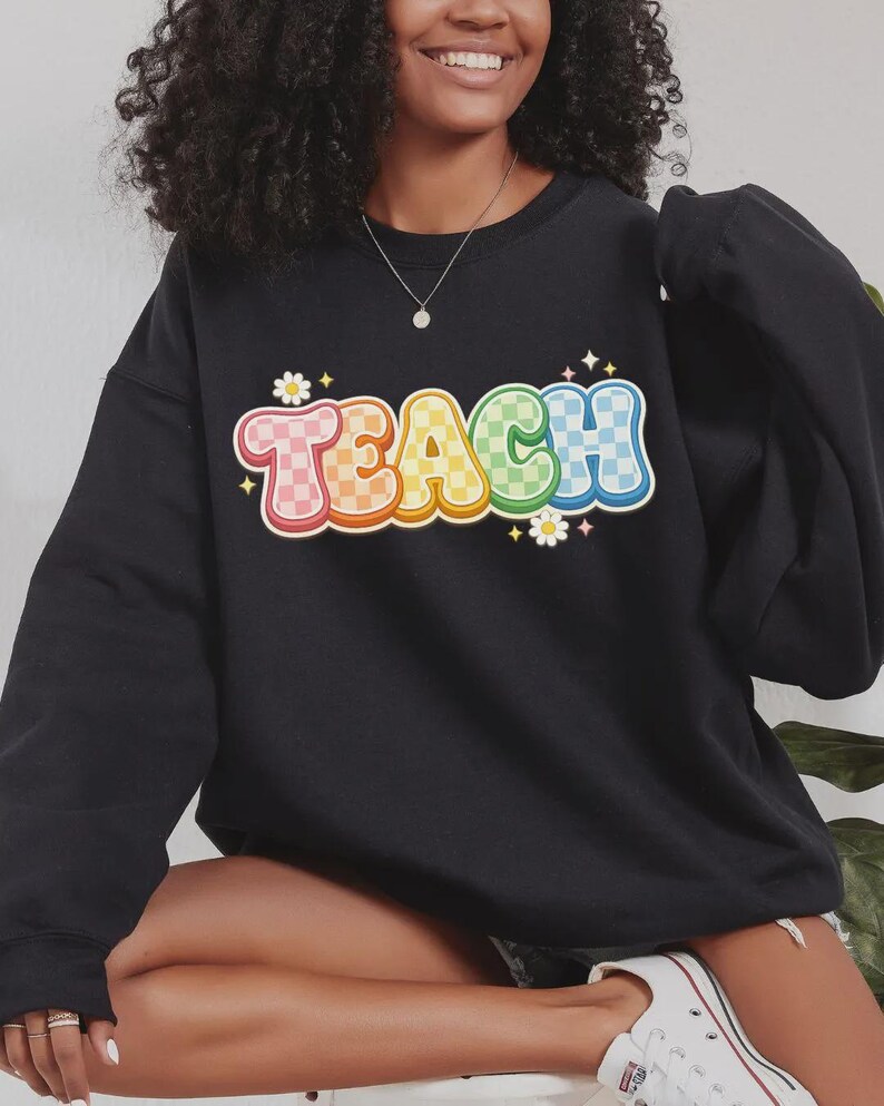 May include: Black sweatshirt with the word "TEACH" in a colorful, retro font. The letters are outlined in white and have a checkered pattern in shades of pink, orange, yellow, green, and blue. Small white flowers and stars accent the design.